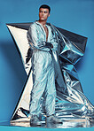 Fashion, cyberpunk and silver with a man on a blue background at a photo shoot in a shiny futuristic outfit. Sci fi, creative or art with a young model in studio for holographic or vaporwave clothes