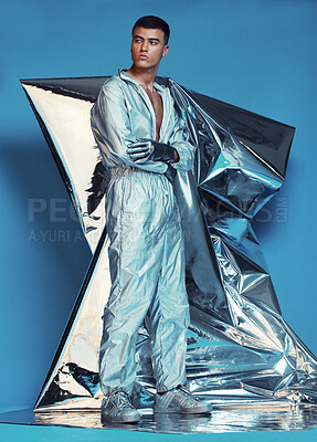 Buy stock photo Fashion, cyberpunk and silver with a man on a blue background at a photo shoot in a shiny futuristic outfit. Sci fi, creative or art with a young model in studio for holographic or vaporwave clothes