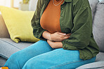 Hands, stomach and menstruation with a woman in pain on a sofa in the living room of her home closeup. Pregnant, gas or indigestion with a female holding her belly for a medical or colon problem