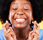 Black woman, face and fingers crossed in studio for hope, trust and optimism of competition, prize and lottery. Happy model emoji for luck, bonus promotion and winning giveaway on yellow background
