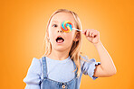 Portrait, cute and a child with a lollipop on a studio background for food, candy or crazy expression. Orange, comic and a girl kid with sweets, treat or sugar for covering an eye on a backdrop