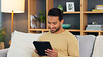 Young man browsing on a digital tablet on a sofa at home. Happy guy smiling and laughing while watching funny videos, scrolling on social media and streaming movies online to enjoy over the weekend