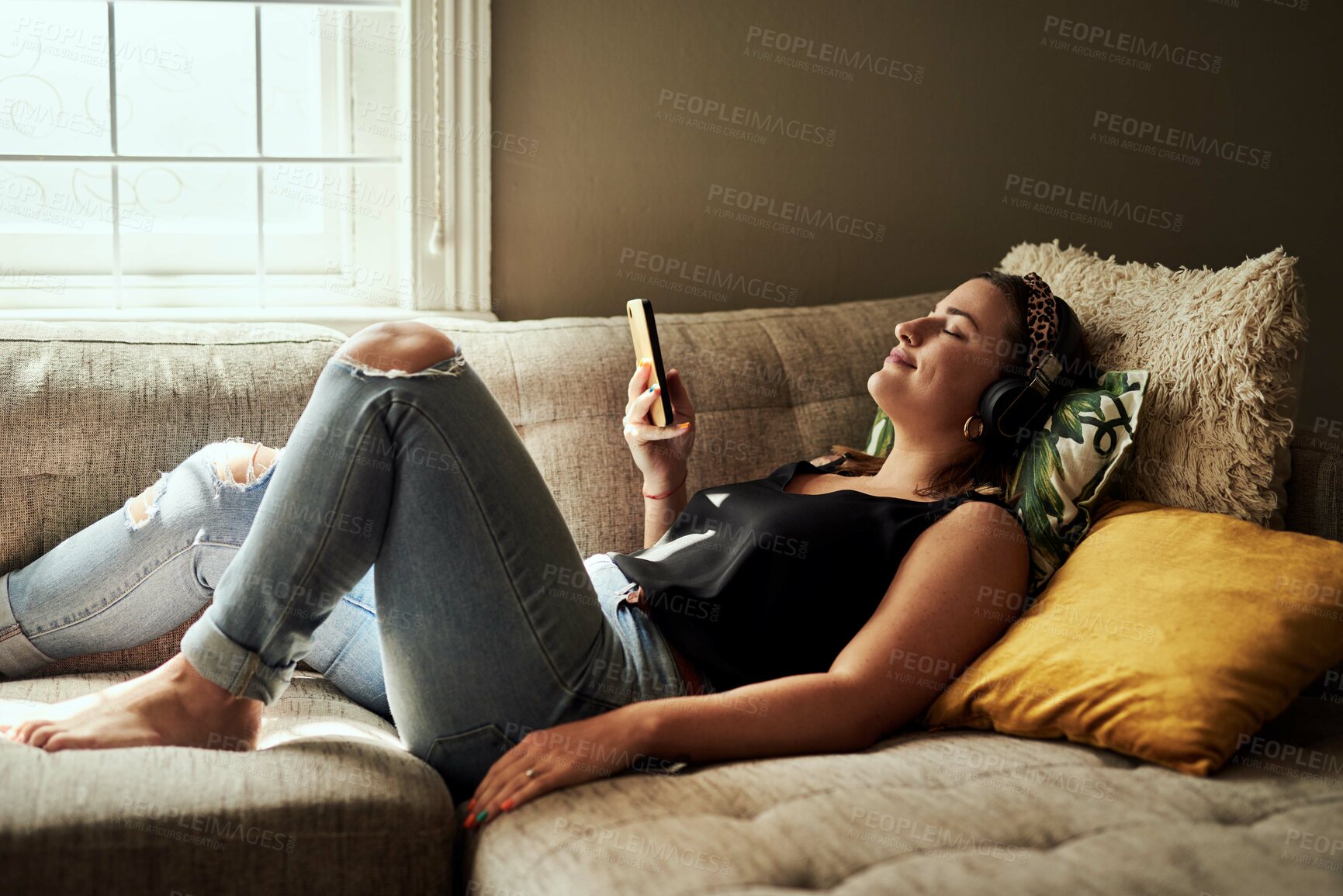 Buy stock photo Shot of a young woman using a smartphone and headphones on the sofa at home
