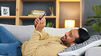 Young man texting on his phone and listening to music on his earphones while lying on the sofa. Happy male browsing social media on an online app and smiling while relaxing on the couch at home