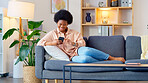 Laughing woman listening to music and sending a message with funny memes on her phone and drinking coffee while on the sofa. Happy black female browsing social media and surfing the internet online
