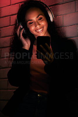 Buy stock photo Woman, phone and headphones for city or night streaming, listening to club music or party in neon light. Happy young person on mobile, social media and audio for techno, rave or network on brick wall