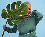 Black woman, monstera leaf and natural beauty, portrait with cosmetics and fashion on blue background. Silk green dress, eco friendly skincare and dermatology, African model glow and plant in studio 