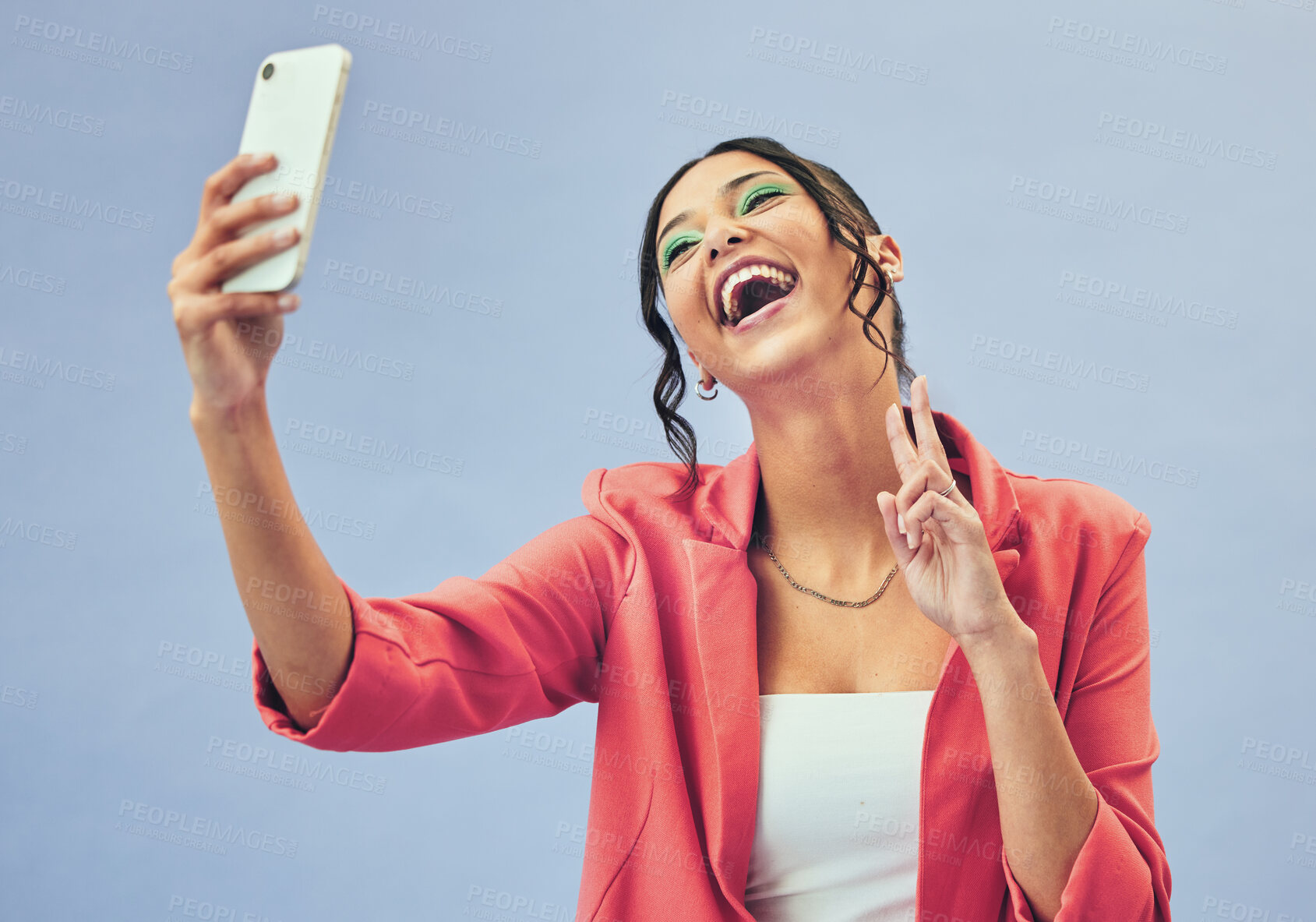 Buy stock photo Phone, laugh selfie and woman with a peace sign from social media and picture for creative job in studio. Influencer, happy and makeup with career fashion with blue background and emoji hand sign