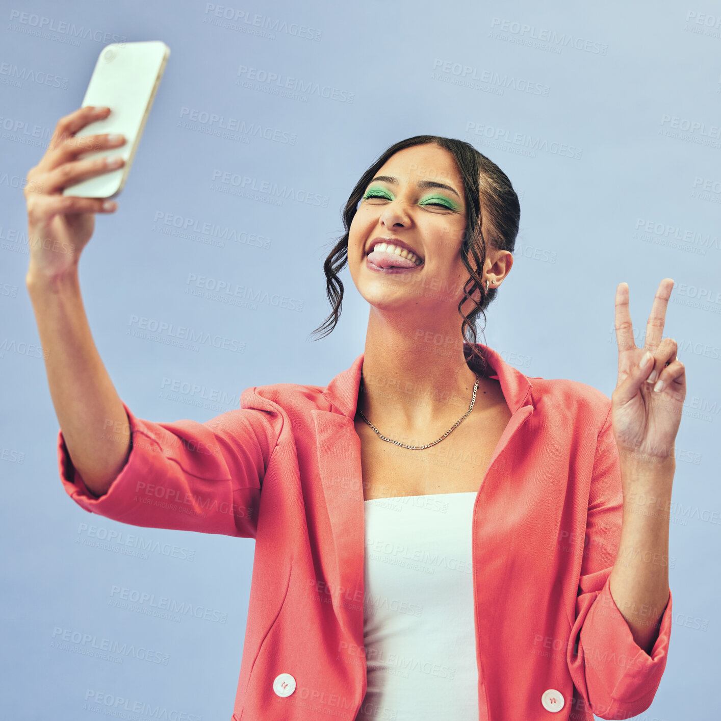 Buy stock photo Phone, fun selfie and woman with a peace sign from social media and tongue out for creative job in studio. Influencer, happy and makeup with career fashion with blue background and emoji hand sign