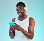 Headphones, phone and black man in a studio networking and listening to music, playlist or album. Happy, smile and African male model scroll on internet with cellphone and song by blue background.