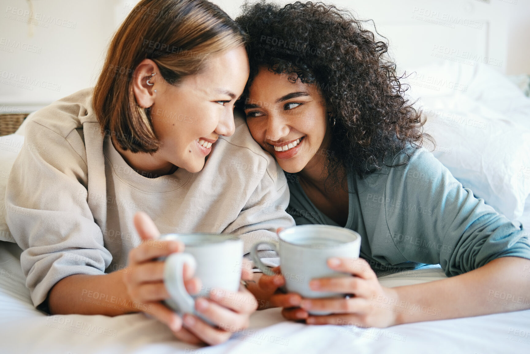 Buy stock photo Coffee, love and lesbian couple on bed in conversation for bonding and relaxing together. Happy, rest and interracial lgbtq women laughing, talking and drinking latte in bedroom of modern apartment.
