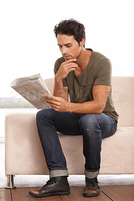 Buy stock photo A handsome young man sitting on the couch at home reading a newspaper