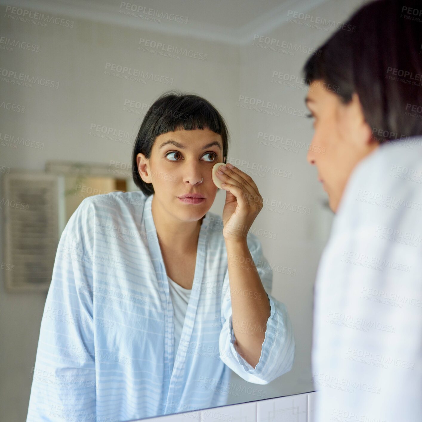 Buy stock photo Shot of a young woman in a bathrobe applying foundation in her bathroom mirror in the morning
