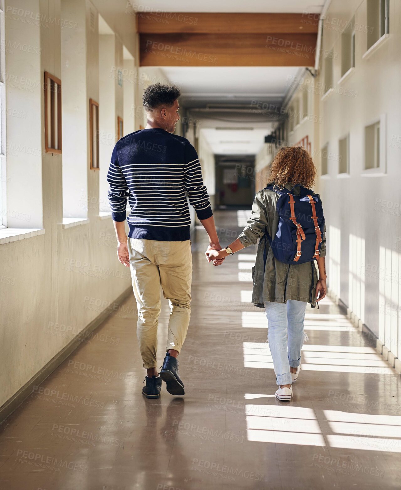 Buy stock photo Rearview shot of an affectionate young couple walking hand in hand through their university corridor