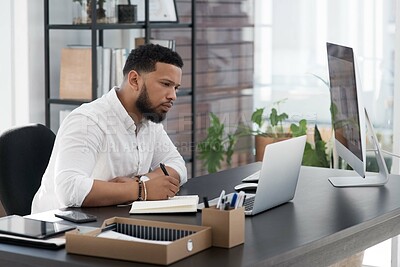 Buy stock photo Shot of a young businessman writing notes while working on a laptop in an office