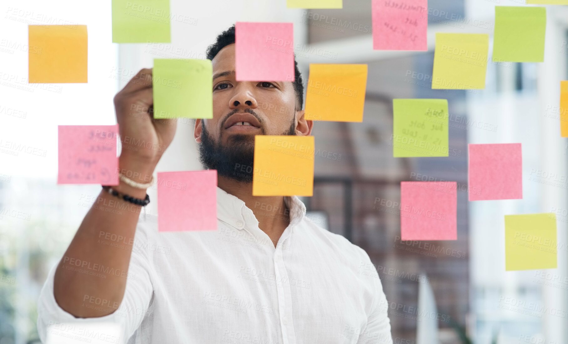 Buy stock photo Shot of a young businessman brainstorming with sticky notes on a glass wall in an office
