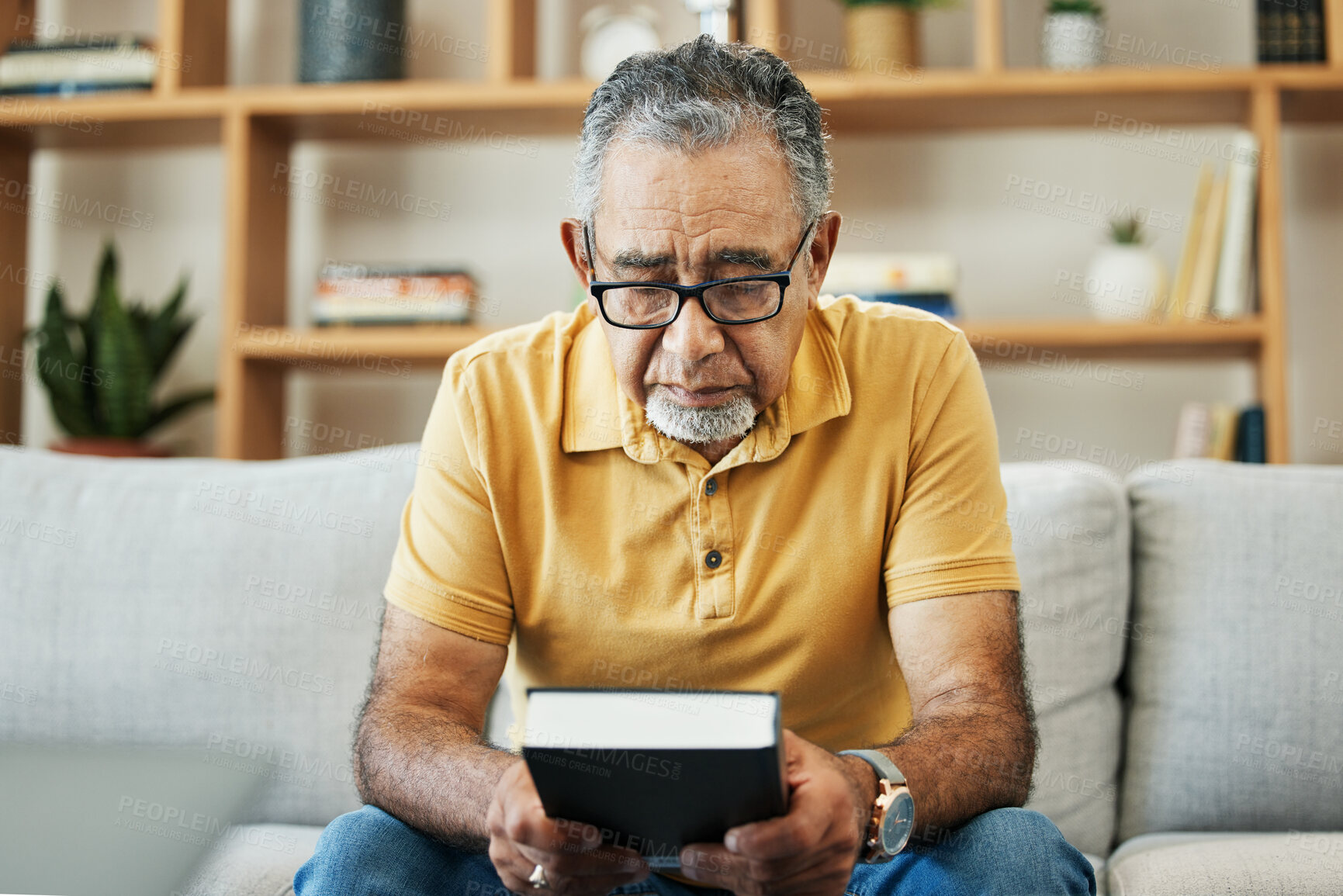 Buy stock photo Mature, man and bible by thinking in home, alone and sadness of memory in living room of thoughts. Elderly, person or grandfather on sofa with hand, holding and book for religion, church or practice