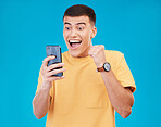 Cellphone, celebration and young man in a studio with fist pump for winning, success or achievement. Happy, smile and male winner from Canada cheering for prize on a phone isolated by blue background