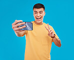 Man, selfie and peace hand sign with happiness, excited with social media with communication on blue background. V emoji, influencer with memory and smile in picture with photography in a studio