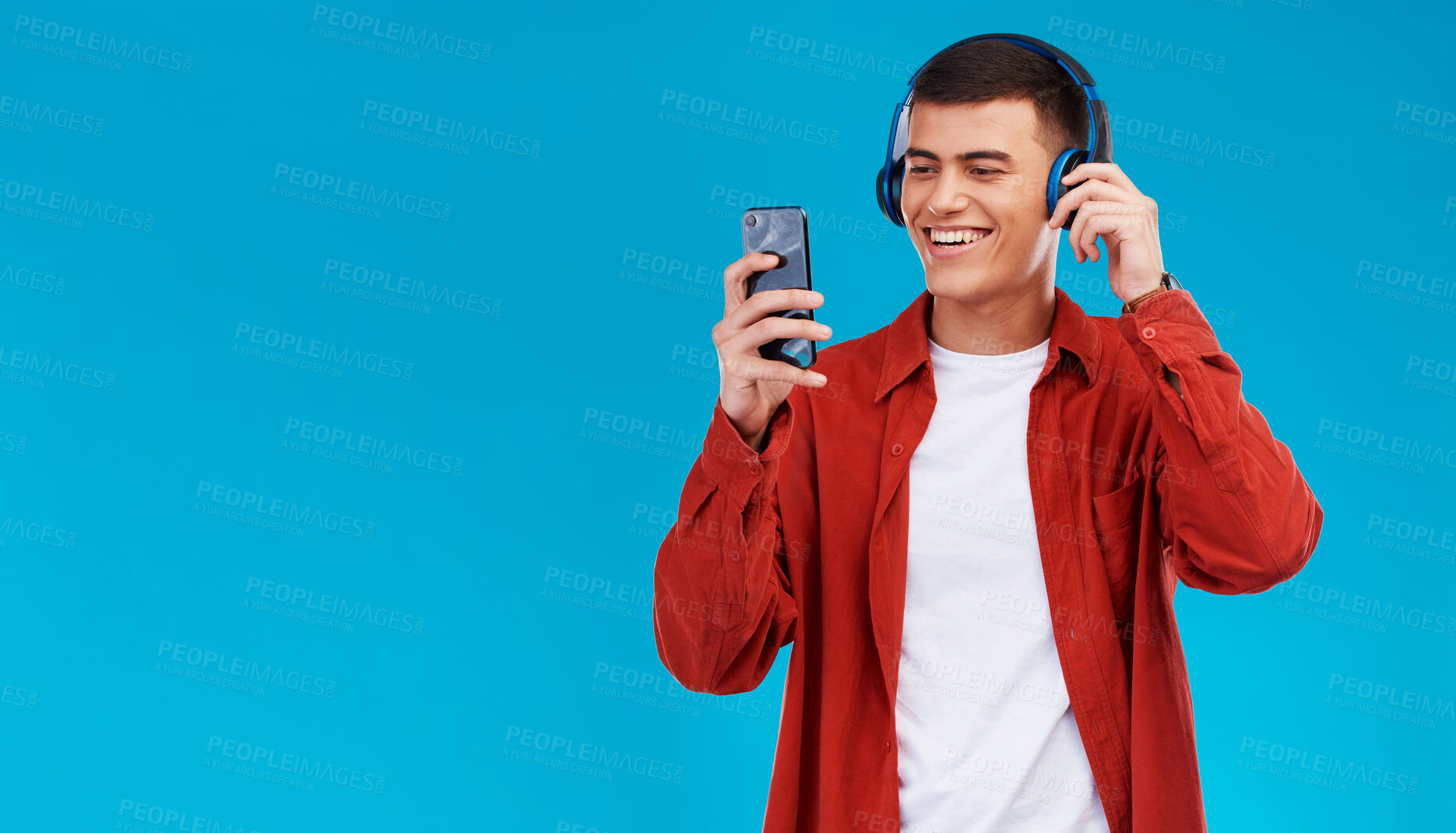 Buy stock photo Man, smile and headphones for music on phone in studio by blue background. Asian person, listening and streaming a song, radio and audio or funny podcast on smartphone, technology and sound for joy