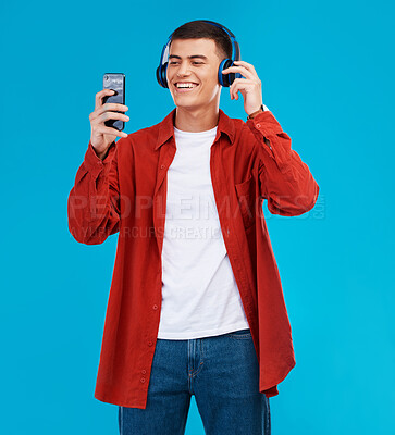 Buy stock photo Man, smile and headphones for audio on phone in studio by blue background. Asian person, searching and streaming a song, radio and music or funny podcast on smartphone, technology and sound for joy