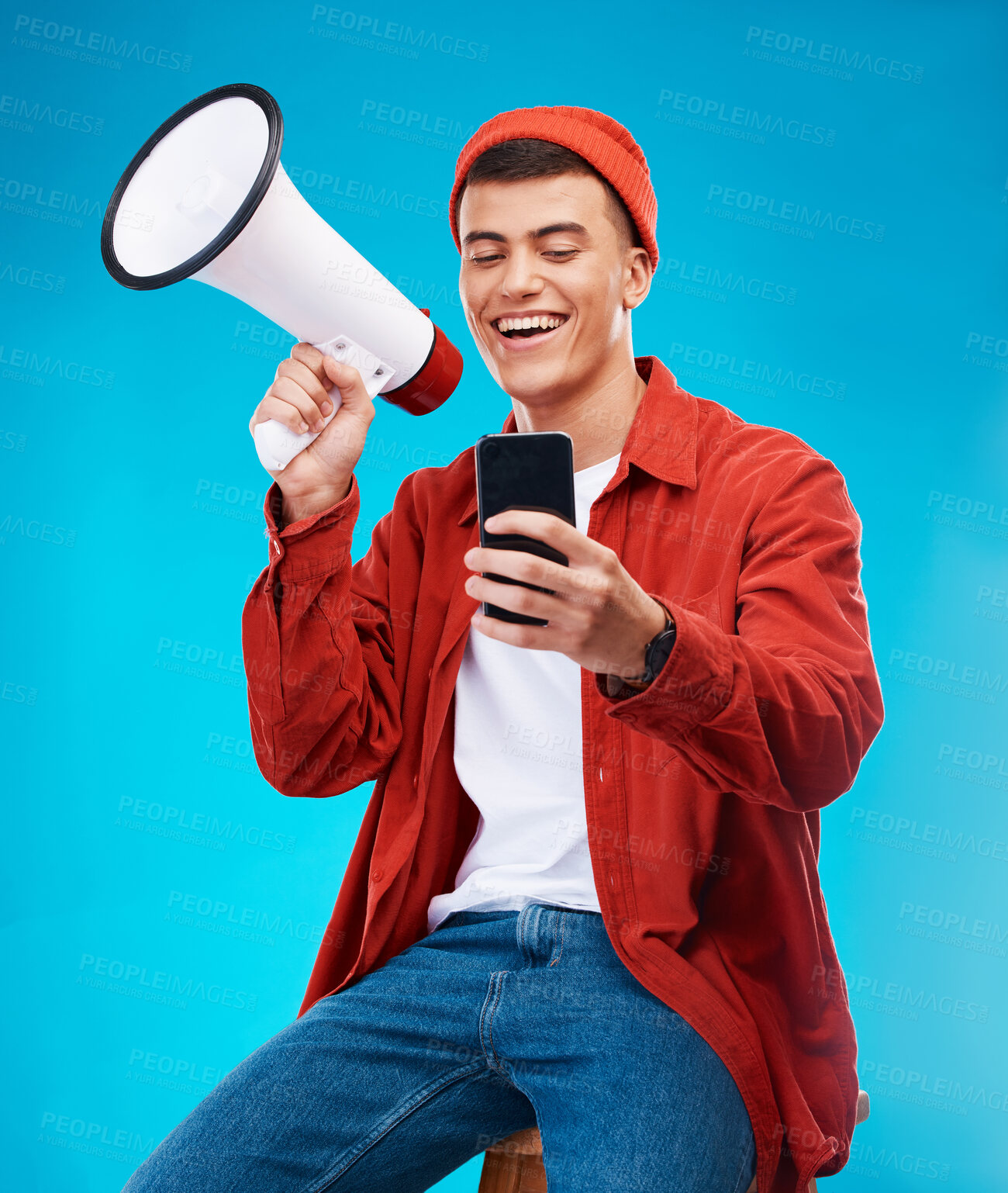 Buy stock photo Phone, megaphone and young man in a studio reading announcement or speech for a rally. Happy, cellphone and male activist or protest with bullhorn for loud communication isolated by blue background