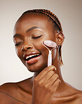 Woman, face roller and massage in studio for skincare, rose quartz cosmetics and aesthetic dermatology on brown background. African model, crystal stone tools and lymphatic drainage for facial beauty