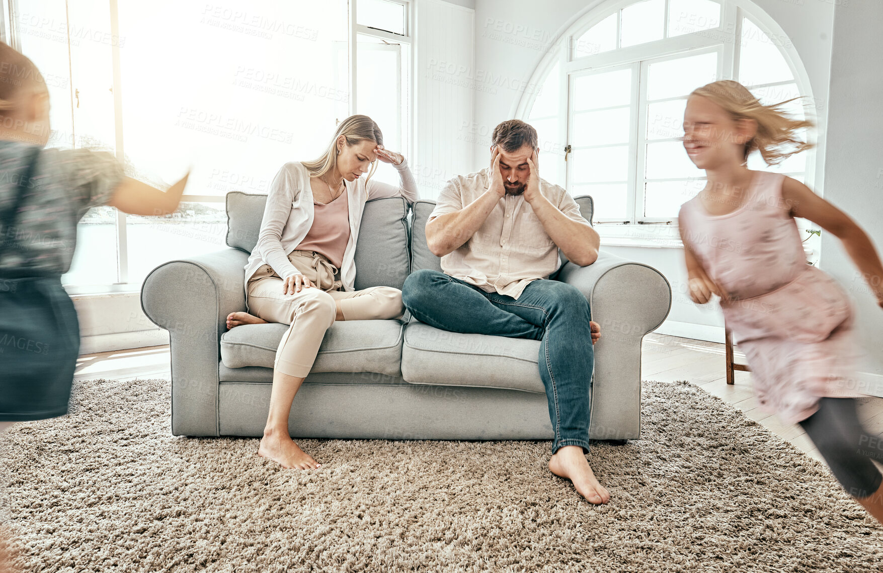 Buy stock photo Frustrated couple, children and running in living room chaos with headache in stress on sofa at crazy or busy home. Parents in burnout with ADHD or hyper active kids or siblings playing wild in house