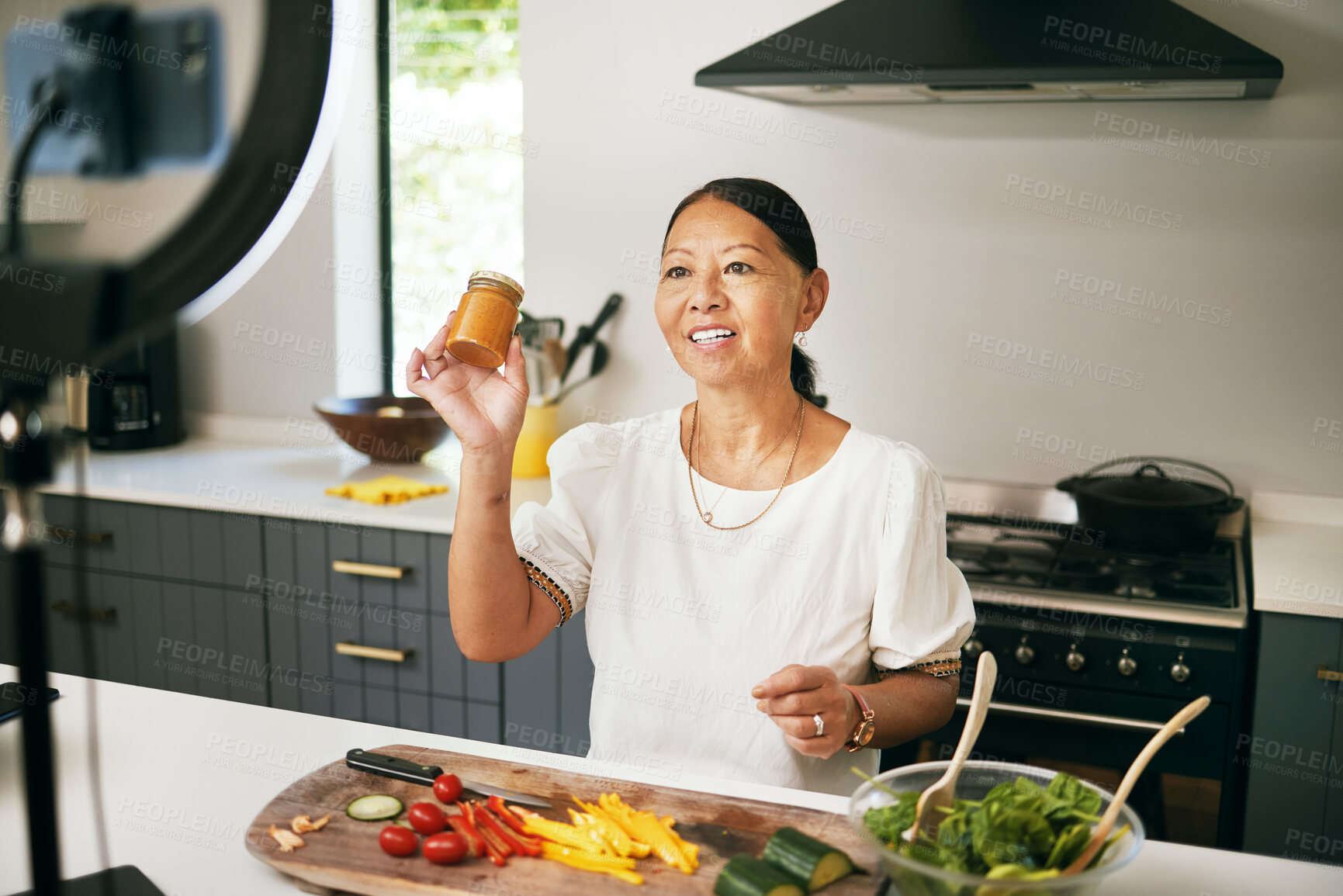 Buy stock photo Asian woman, happy and live streaming for cooking with bottle, healthy food and social media blog in kitchen. Chef, mature influencer and vegetables for content creation, broadcast tutorial and phone