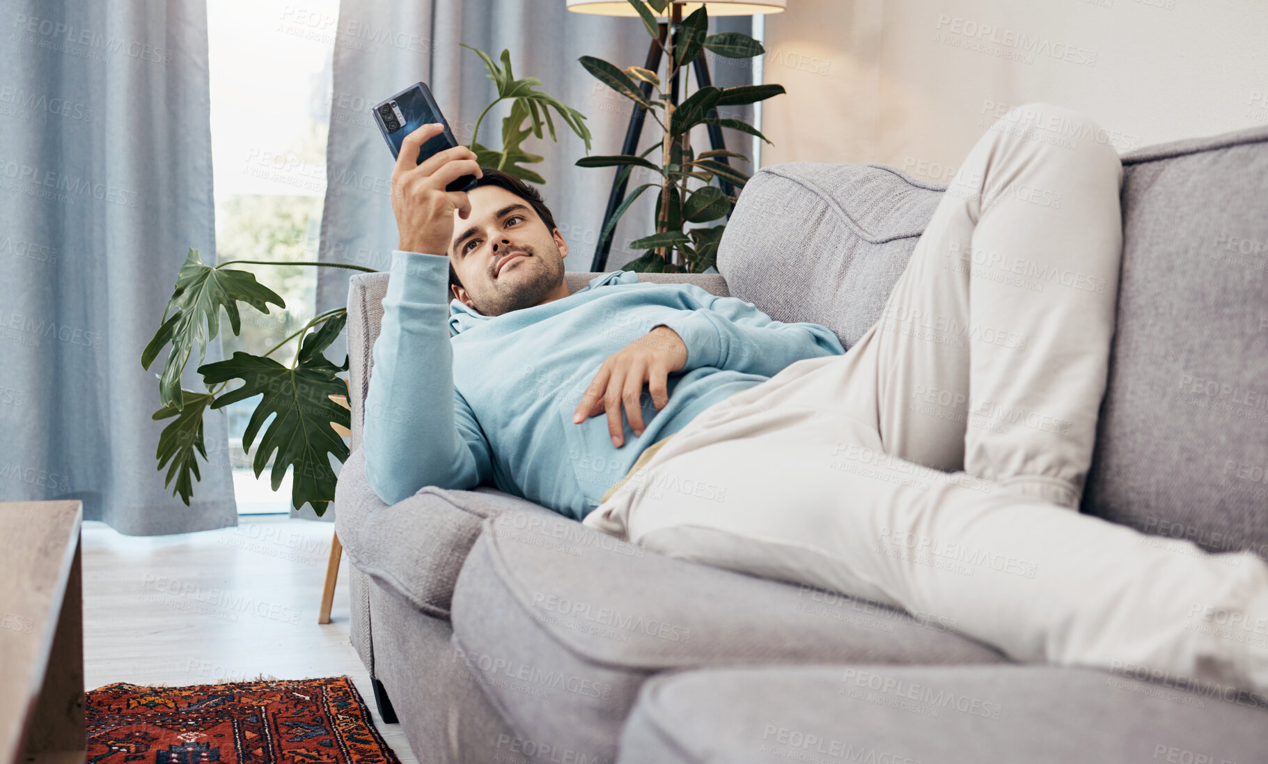 Buy stock photo Man, phone and relax on sofa for social media, communication or entertainment in living room at home. Male person lying on lounge couch with mobile smartphone for online app or streaming at house