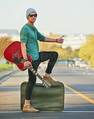 Buy stock photo Shot of a handsome young man hitchhiking at the side of the road with a guitar and suitcase
