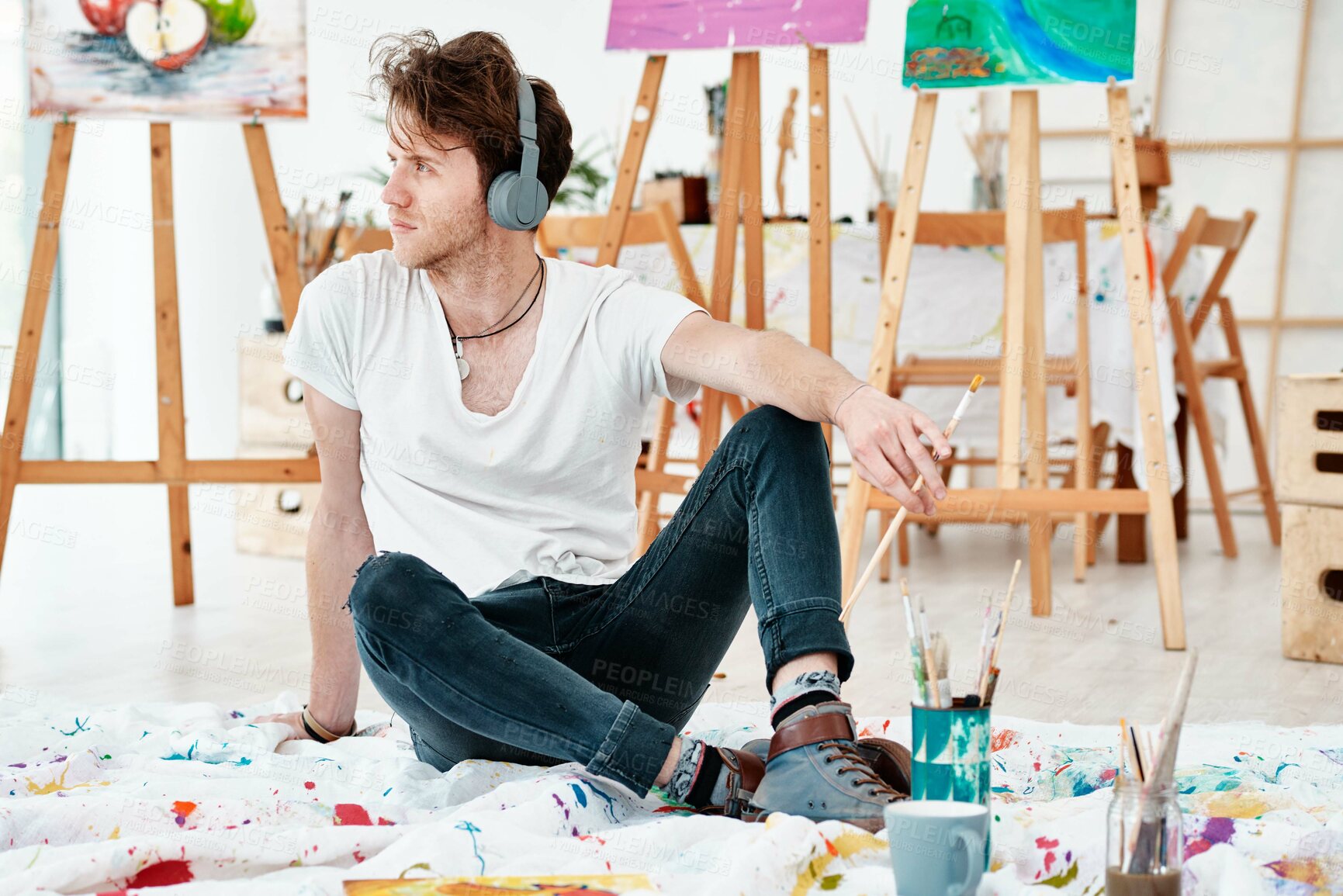 Buy stock photo Full length shot of a handsome young artist sitting alone and looking contemplative while listening to music through headphones
