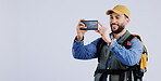 Hiking, backpack and happy man with phone in studio for video call, photography or memory on grey background. Travel, smartphone and male backpacker with app for live streaming journey in nature