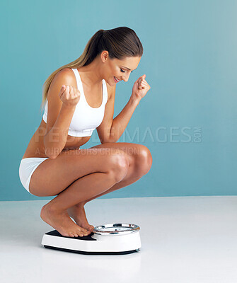 Buy stock photo Studio shot of a fit young woman weighing herself on a scale and cheering against a blue background