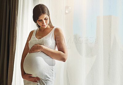 Buy stock photo Shot of a pregnant woman cradling her belly while standing in front of a window at home