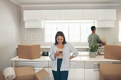 Buy stock photo Shot of a woman using her cellphone while her partner unpacks boxes in their new home