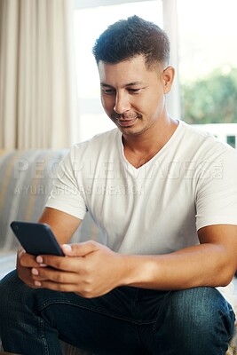 Buy stock photo Shot of a handsome young man sitting alone on the sofa at home and using his cellphone