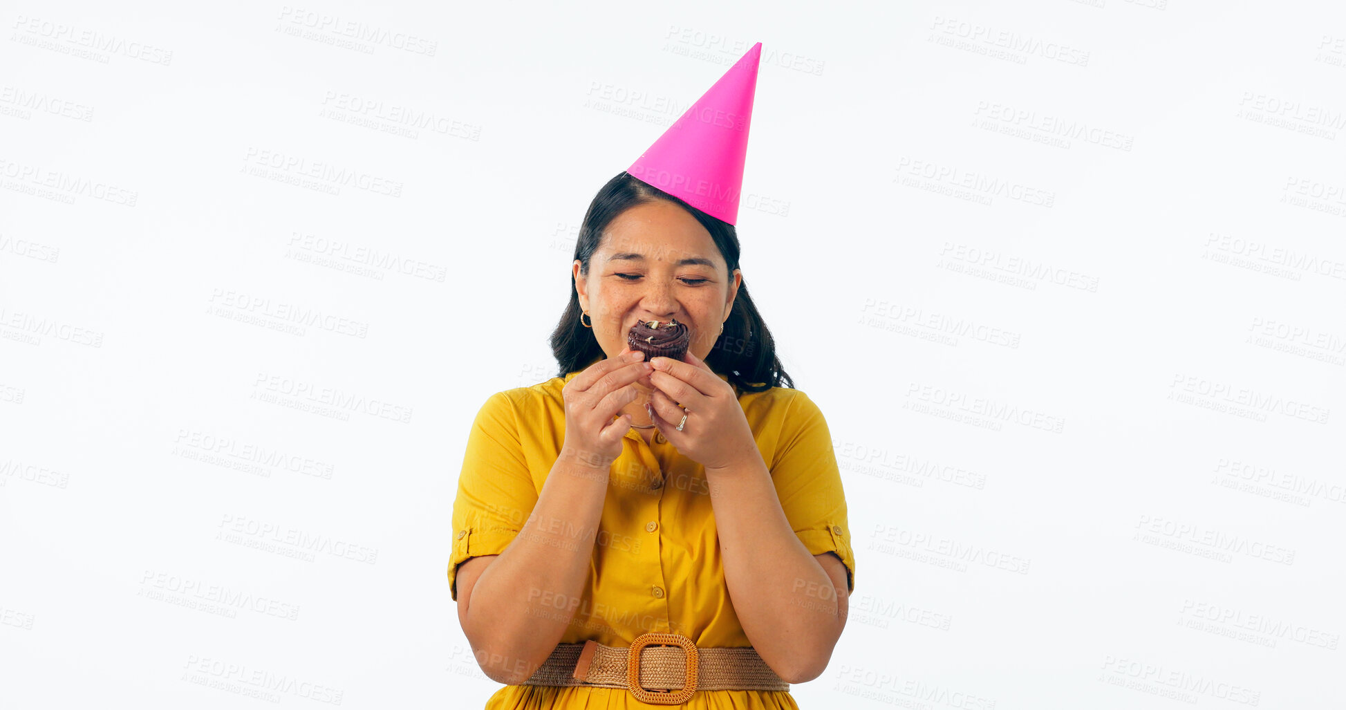 Buy stock photo Birthday, party and an asian woman eating a cupcake in studio isolated on a white background for celebration. Smile, hat and a happy young person with a chocolate dessert or snack at an event