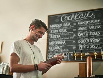 Buy stock photo Shot of a happy young bartender holding a bottle of liquor while standing behind the bar
