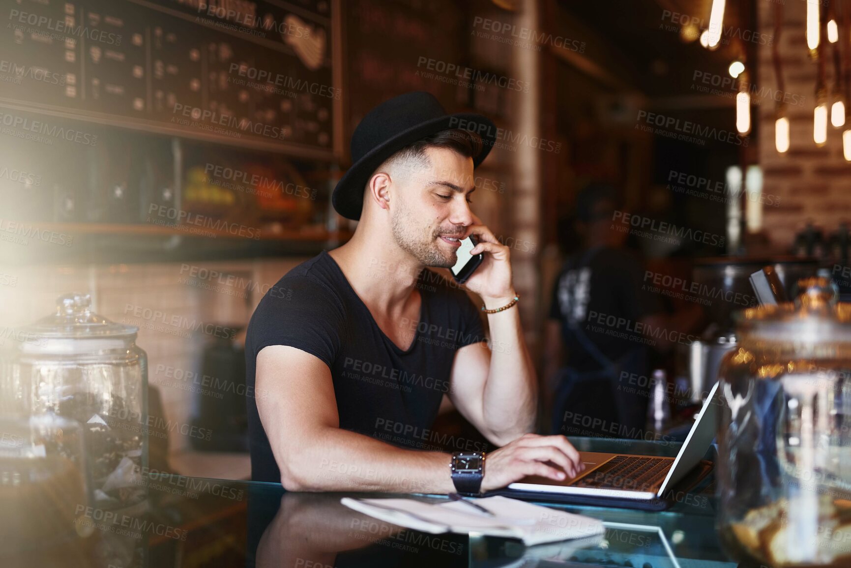 Buy stock photo Shot of a young man using a phone and laptop while working in a coffee shop