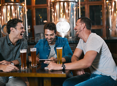Buy stock photo Shot of three cheerful young men drinking beer together at a table inside of a beer brewery during the day