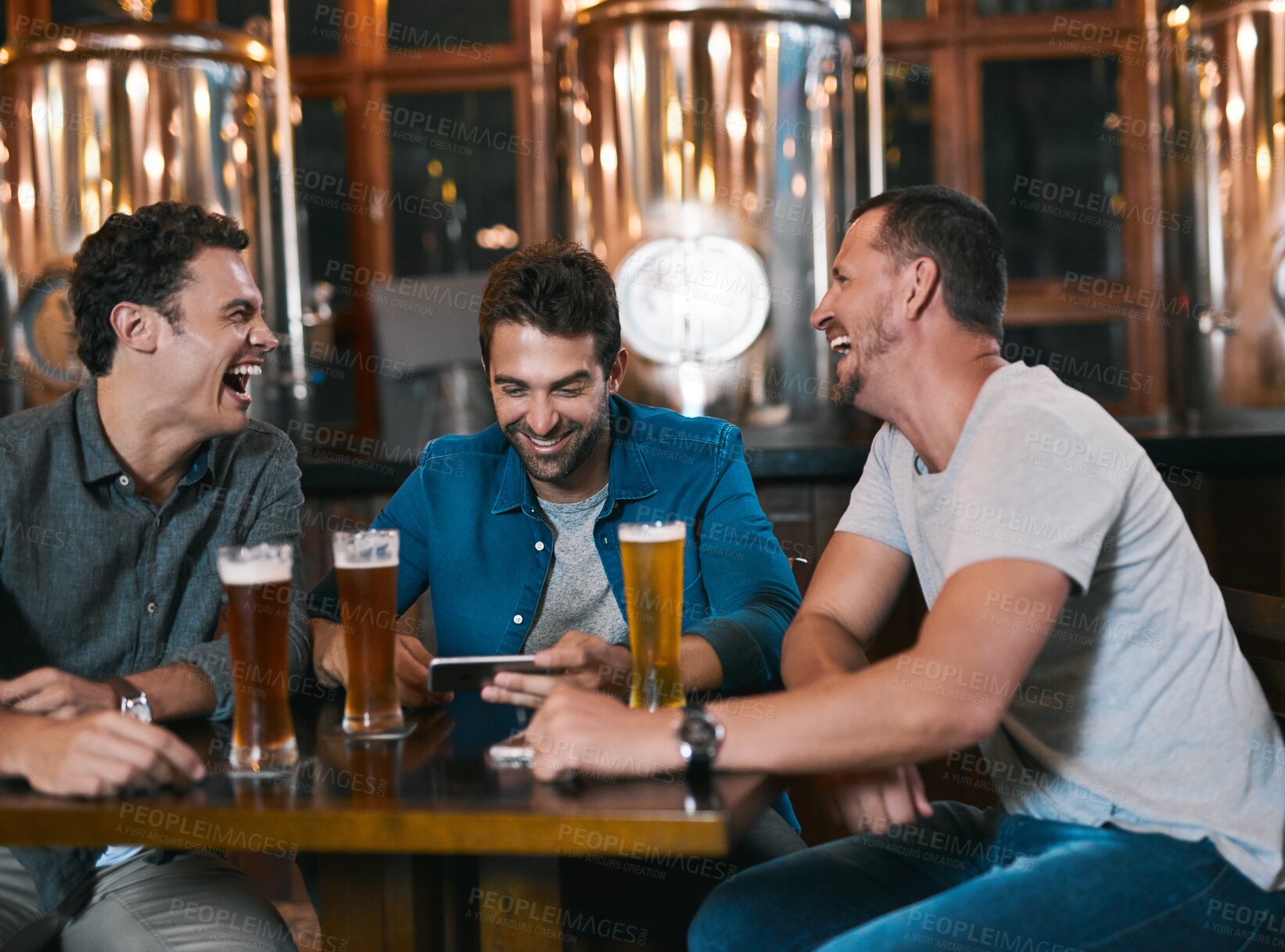 Buy stock photo Shot of three cheerful young men drinking beer together at a table inside of a beer brewery during the day