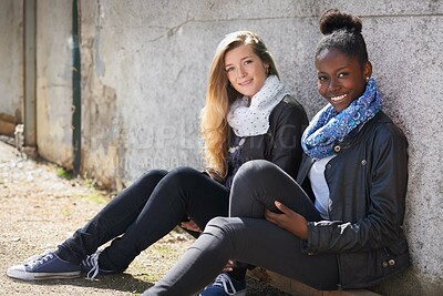 Buy stock photo Stylish, women sitting outdoor at wall at campus and smiling. Young female friends, happy diverse students enjoying bonding or quality time and trendy or fashion and having fun on their day off