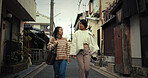 Japanese women, friends and walk in city, talking and travel together on vacation, street and buildings. People, girl and outdoor in metro road, houses or urban neighborhood in conversation in Tokyo