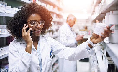 Buy stock photo Shot of a cheerful young female pharmacist talking on her cellphone while reading the back of a medication box