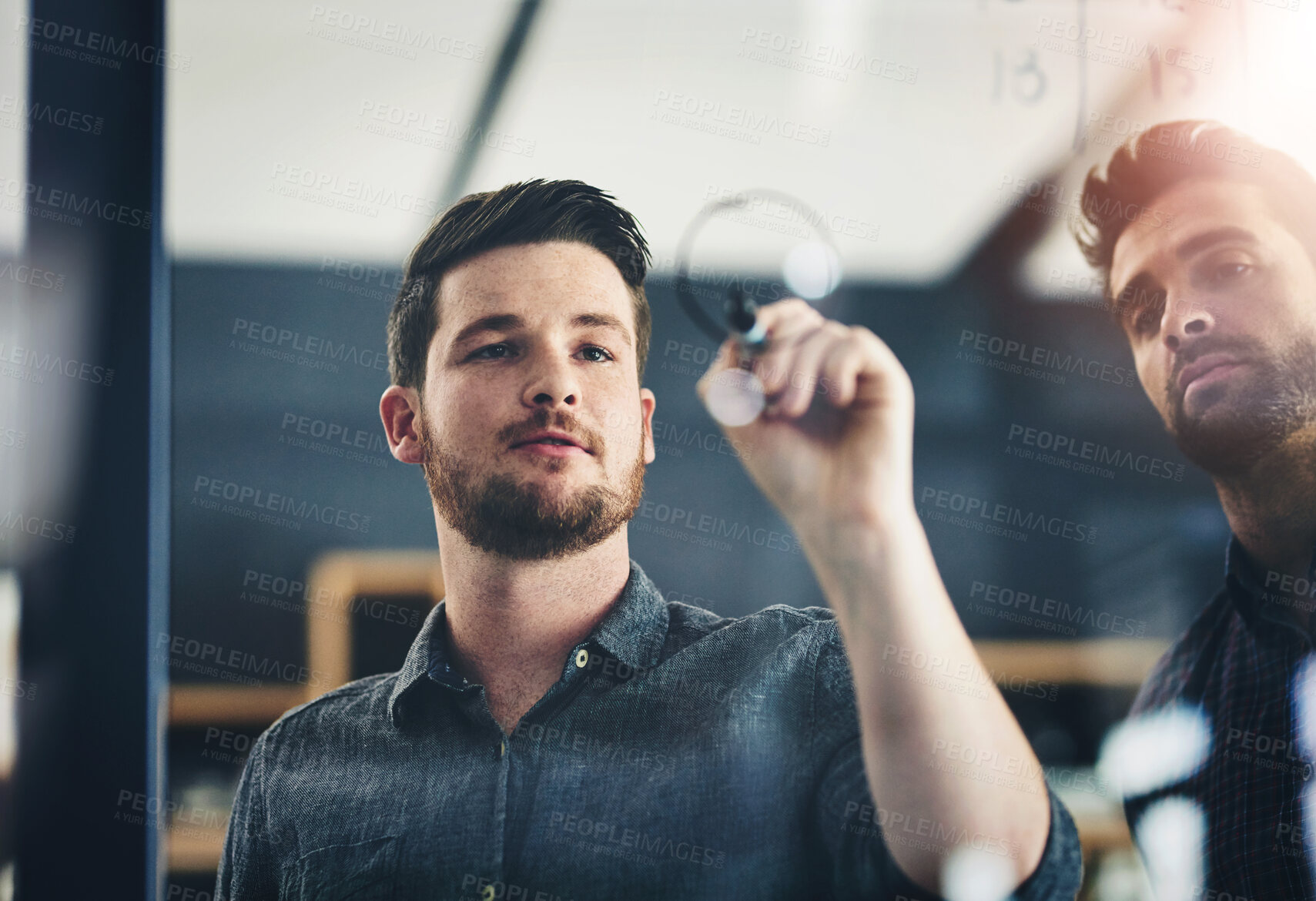 Buy stock photo Shot of two young businessmen brainstorming on a glass wall in an office