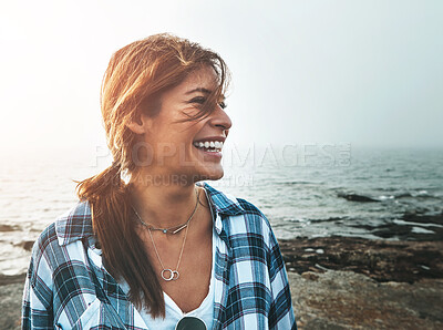 Buy stock photo Shot of a cheerful young woman standing on rocks next to the ocean while laughing outside during the day