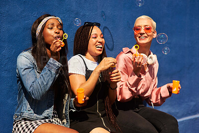 Buy stock photo Cropped shot of three attractive young women sitting against a blue wall together and bonding by blowing bubbles
