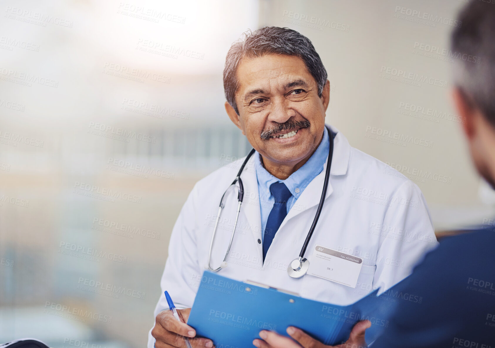 Buy stock photo Shot of a cheerful mature male doctor seated at his desk while consulting a patient inside a hospital during the day