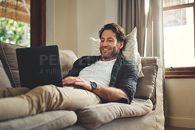 Buy stock photo Shot of a handsome young man using his laptop while relaxing on a couch at home